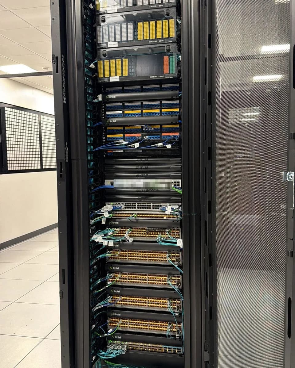 Open datacenter rack on the floor: Corning fiber panels up top, copper panels and switches below, aqua patch cords routed neatly down the side.